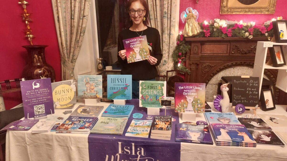 Author Isla Wynter standing behind a table full of her books at an event