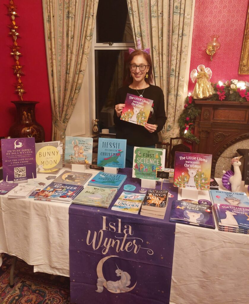Author Isla Wynter standing behind a table full of her books at an event